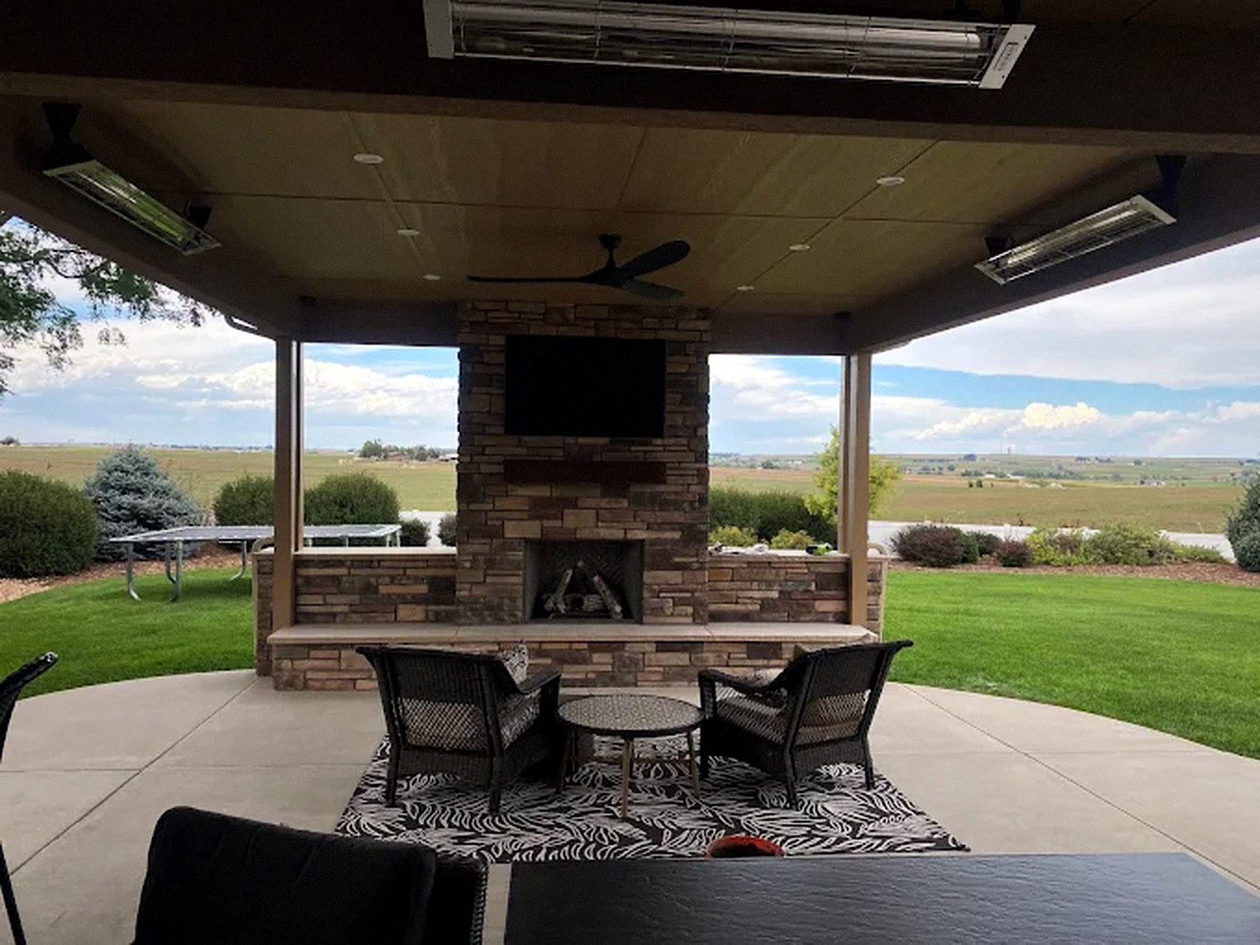 Covered concrete patio with a stone fireplace, ceiling fan, infrared heaters and lounge seating in Parker, CO